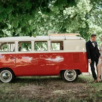 Brautpaar lehnt an einem rot-weißen VW-Bus unter Bäumen