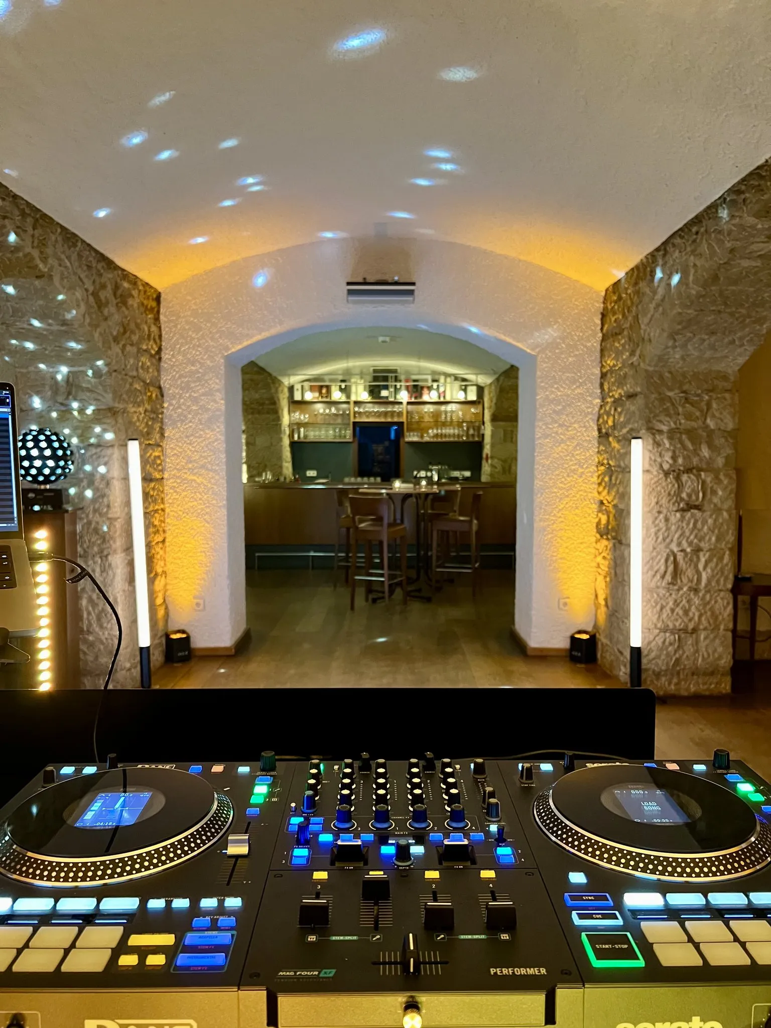 Blick über einen DJ-Controller durch einen Gewölbekeller, an den Seiten leuchtende Lichtstelen, hinten durch einen Steinbogen eine beleuchtete Bar mit einem Regal voller Gläser