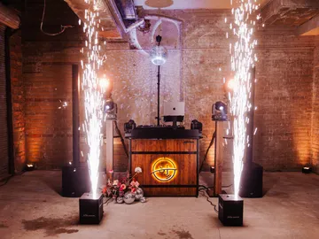 DJ-Pult mit leuchtendem SJ-Logo zwischen zwei Funkenfontänen in einem Saal mit Backsteinwänden, darüber eine Discokugel