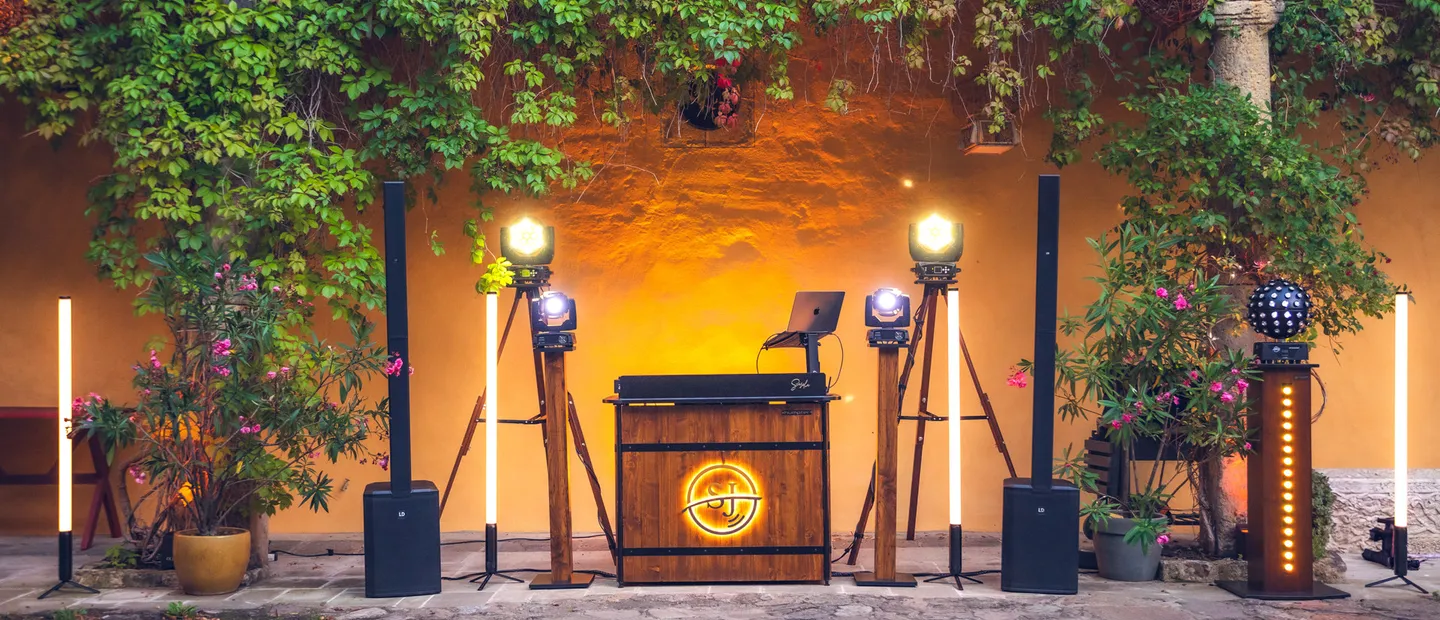 Beleuchtetes DJ-Pult aus Holz mit SJ-Logo, Lautsprechern und Lichttechnik in einem Innenhof mit Kletterpflanzen