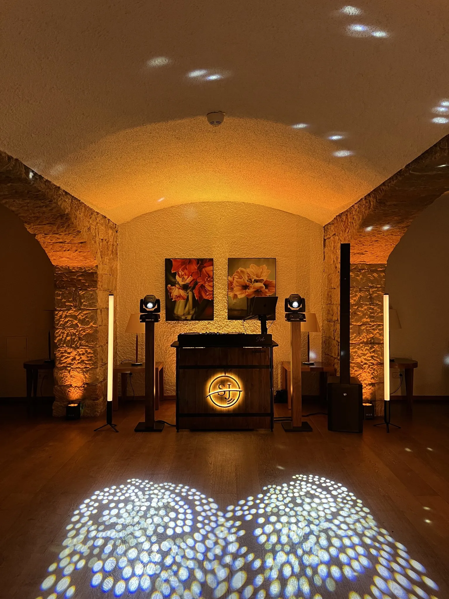 DJ-Pult mit leuchtendem Logo, Lichtstelen und Scheinwerfern in einem Gewölbekeller, warmes Licht, Lichtmuster auf dem Holzboden, zwei Bilder an der Stirnwand