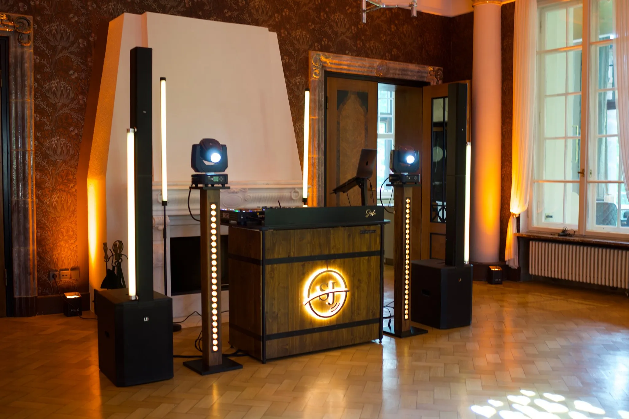 Holzverkleidetes DJ-Pult mit beleuchtetem Monogramm, Lichtstelen und zwei Scheinwerfern vor einem hellen Kamin auf Fischgrätparkett