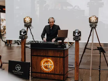 DJ Sascha Juranek mit Kopfhörern an seinem Holzpult mit SJ-Logo, daneben Scheinwerfer, links eine Gitarre und ein Koffer mit seinem Namen