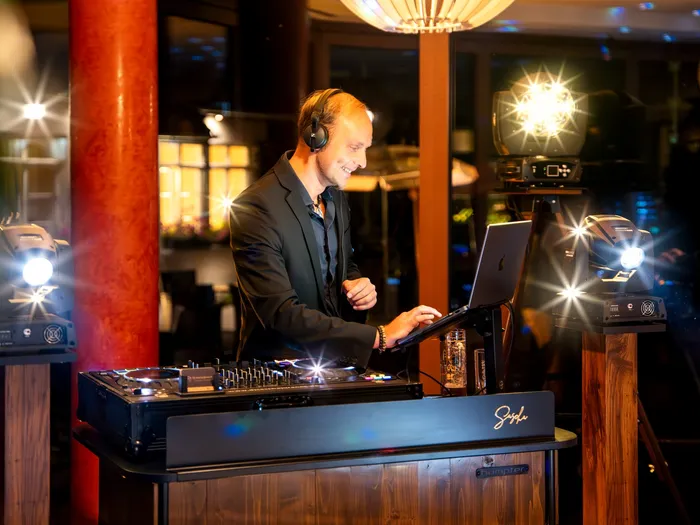 DJ Sascha Juranek lacht mit Kopfhörern an seinem DJ-Pult, um ihn leuchten Scheinwerfer auf Holzsäulen
