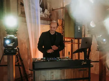 DJ Sascha Juranek lächelt hinter seinem DJ-Pult, links ein hell strahlender Scheinwerfer und ein heller Vorhang