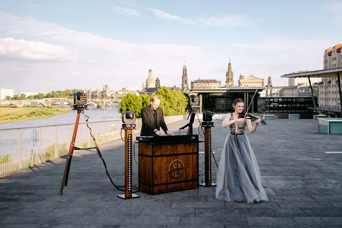 DJ Sascha Juranek am Holzpult und eine Geigerin auf einer Terrasse über der Elbe, dahinter die Dresdner Altstadt mit der Frauenkirche