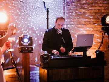 DJ Sascha Juranek am Pult, Kopfhörer um den Hals, hinter ihm eine Backsteinwand mit Lichtpunkten einer Discokugel