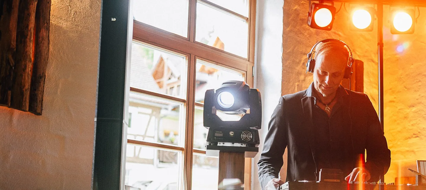 DJ Sascha Juranek mit Kopfhörern am Pult, über ihm orange leuchtende Strahler, links ein hohes Sprossenfenster