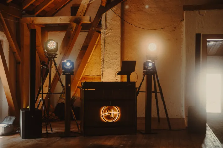 DJ-Pult mit leuchtendem SJ-Logo, daneben Lichttechnik auf Stativen unter einem Dachstuhl