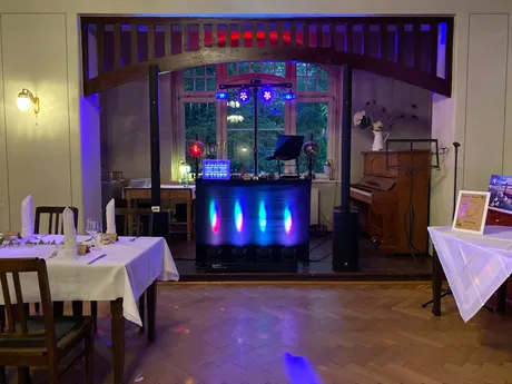 DJ-Pult mit Lichteffekten unter einem Holzbogen im Fischhaus, daneben ein Klavier und gedeckte Tische