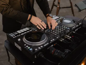 Hände mit Ringen und Holzarmbanduhr an einem DJ-Controller, daneben eine Kamera im Pult