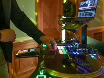 Eine Hand am Mischpult mit bunt leuchtenden Tasten, dahinter ein Laptop mit DJ-Software