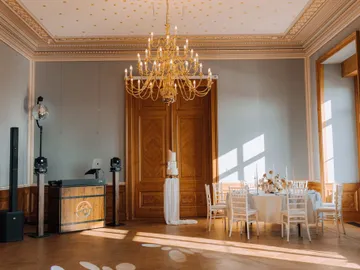 DJ-Pult mit Anlage und Discokugel in einem Festsaal mit Kronleuchter, daneben eine gedeckte Tafel und die Hochzeitstorte