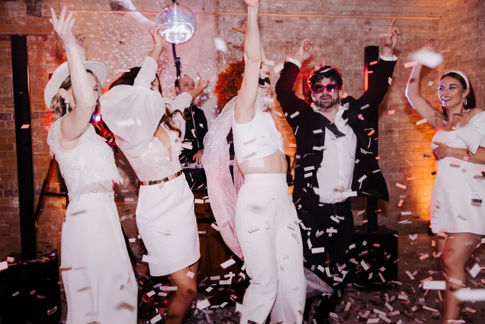 Hochzeitsgäste tanzen im Konfettiregen unter einer Discokugel, dahinter der DJ am Pult