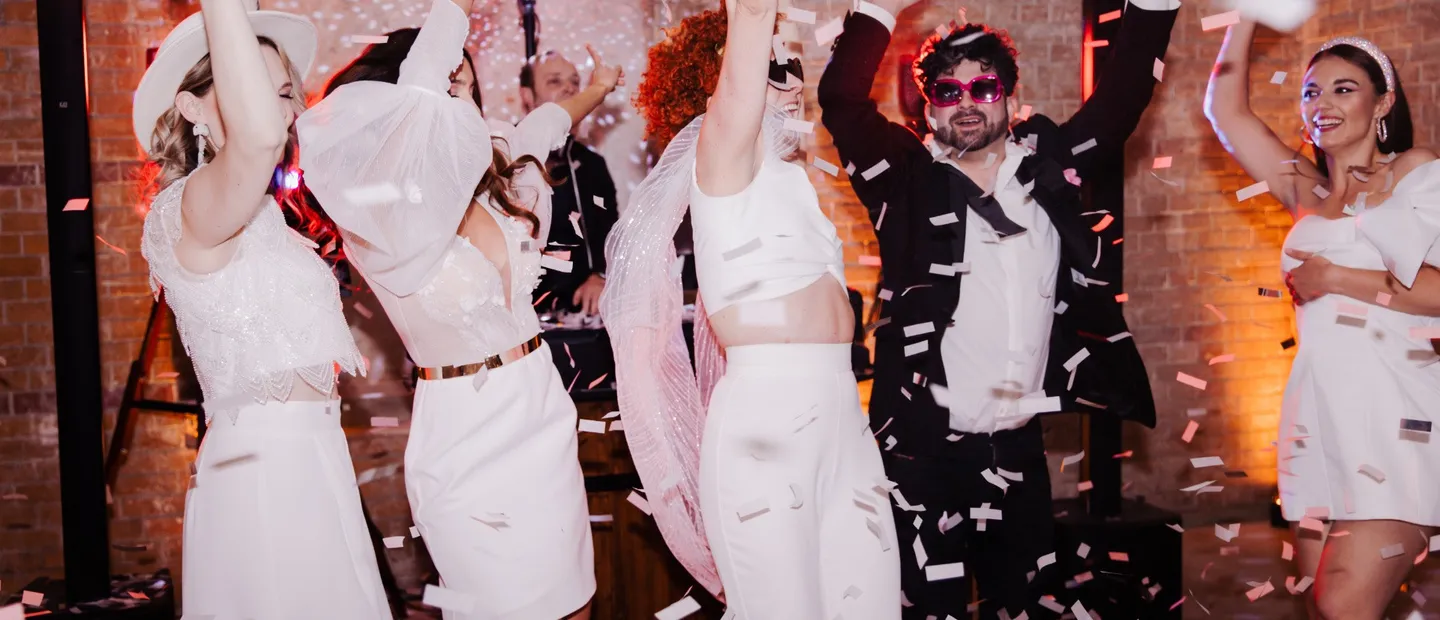 Hochzeitsgäste tanzen im Konfettiregen unter einer Discokugel, dahinter der DJ am Pult