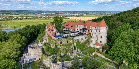 Schloss Scharfenberg von oben: die Anlage mit roten Ziegeldächern, Rundturm und Ringmauer auf einem bewaldeten Hang, dahinter Felder, ein Ort und ein Gewässer im Tal