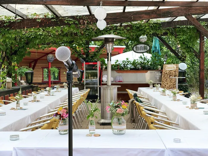 Mikrofon auf einem Ständer vor langen, weiß gedeckten Tafeln mit Blumen unter einer bewachsenen Pergola
