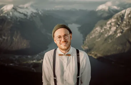 Sebastian mit Mütze, Brille und Holzfliege vor Bergpanorama
