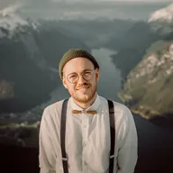 Sebastian mit Mütze, Brille und Holzfliege vor Bergpanorama