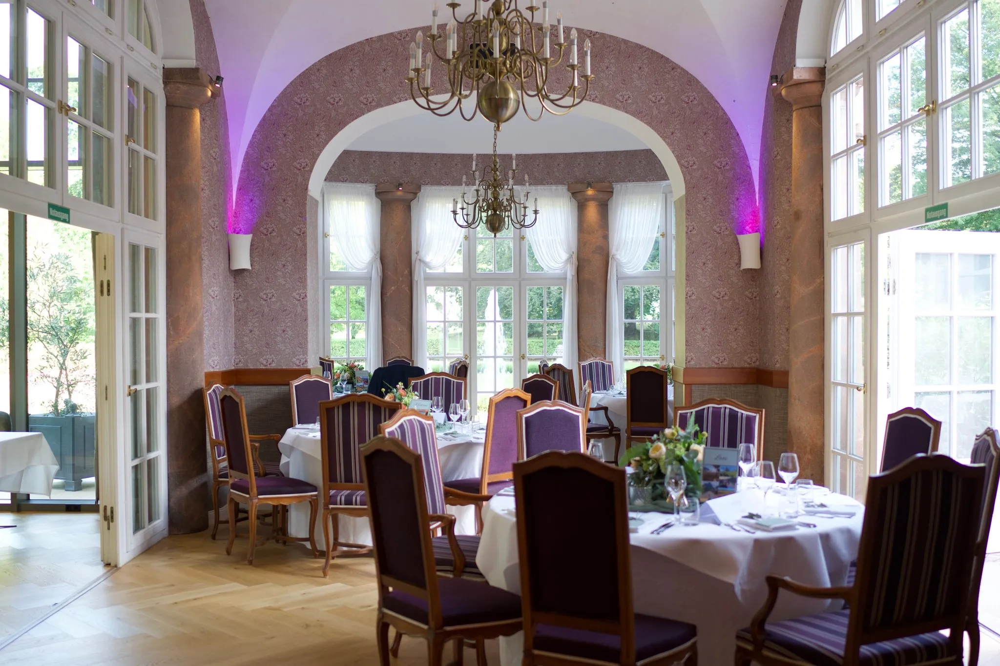 Saal mit Gewölbebogen und zwei Messingkronleuchtern, runde Tische mit weißen Decken und violett gepolsterte Stühle
