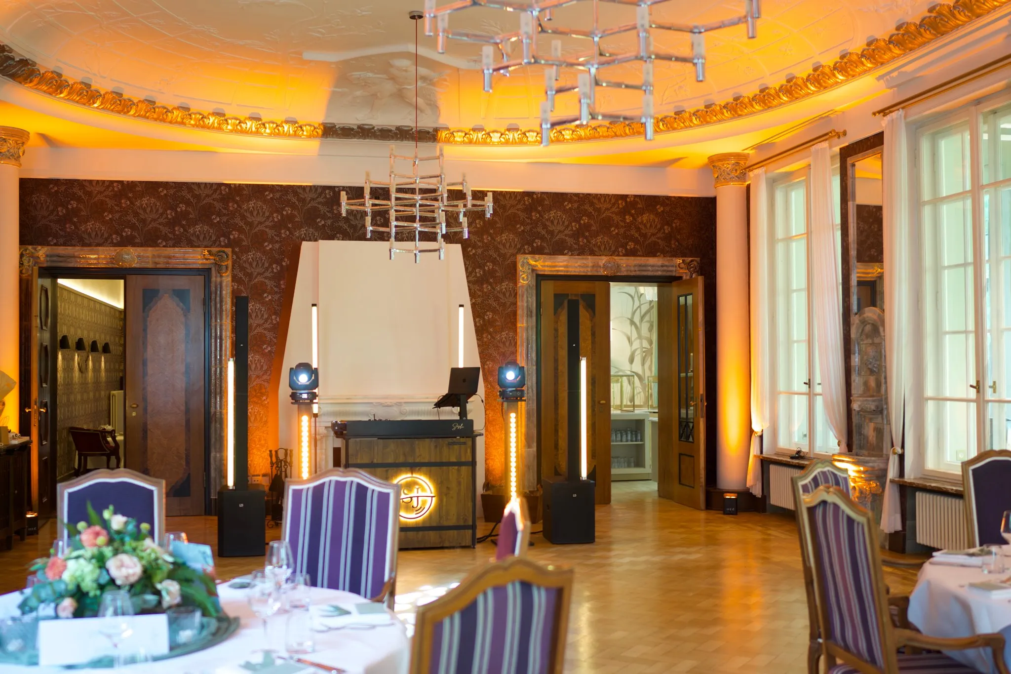 Saal mit vergoldetem Deckenstuck und Kronleuchtern, im Hintergrund ein beleuchtetes DJ-Pult, vorn eingedeckte Tische mit Blumenschmuck