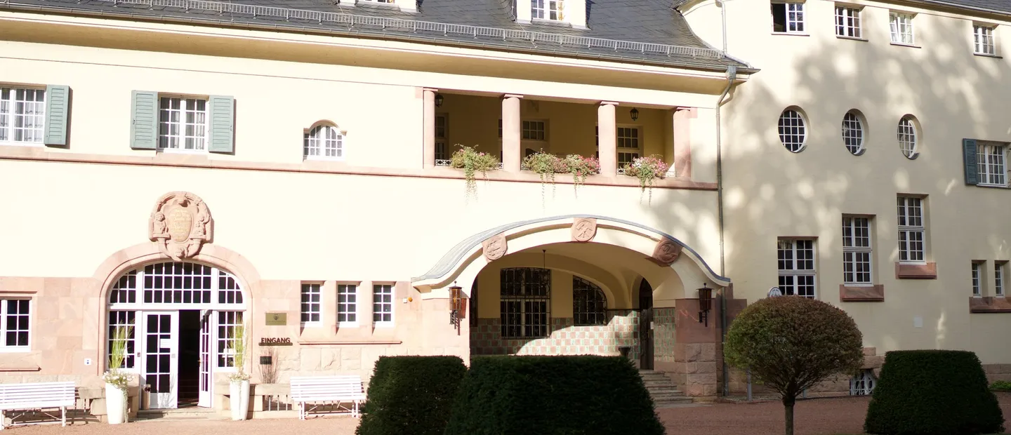 Cremefarbene Schlossfassade mit Mansarddach, Rundbogenportal und offener Loggia, davor geschnittene Hecken und weiße Bänke auf dem Kiesvorplatz