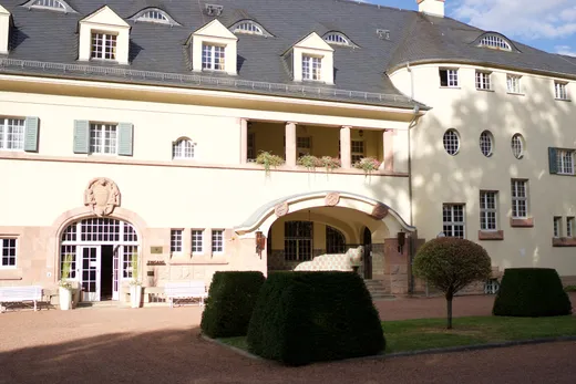 Cremefarbene Schlossfassade mit Mansarddach, Rundbogenportal und offener Loggia, davor geschnittene Hecken und weiße Bänke auf dem Kiesvorplatz