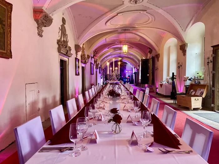 Lange, weiß gedeckte Tafel mit roten Servietten, Rosen und Kerzenleuchter in einem Schlossgang mit Gewölbedecke und violettem Licht
