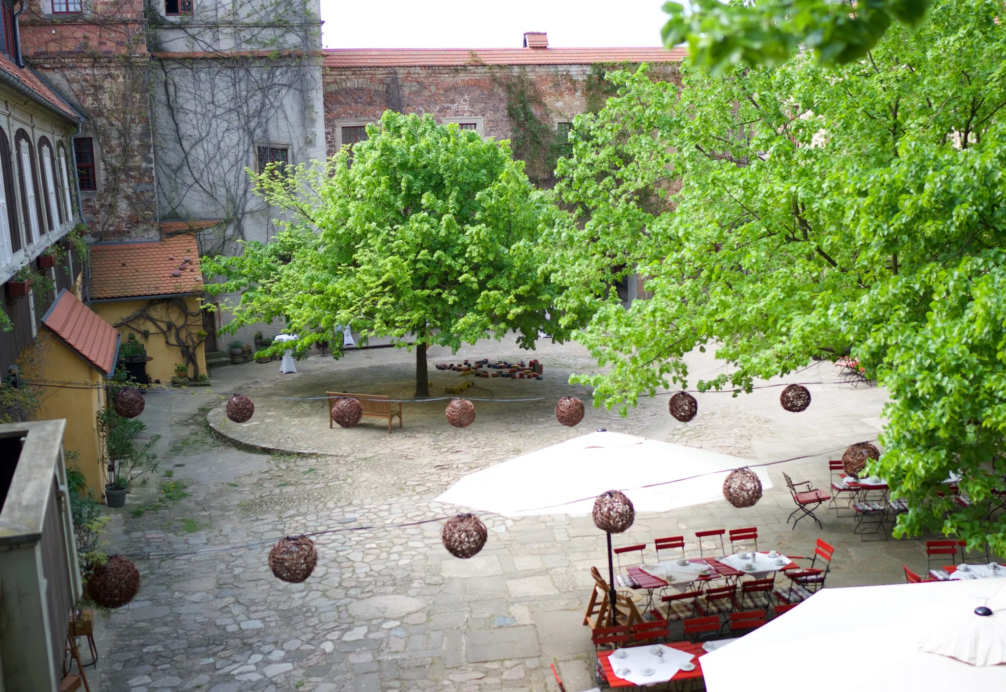 Blick von der Galerie in den gepflasterten Schlosshof mit großem Baum, Lampionschnur, weißem Sonnenschirm und eingedeckten roten Tischen