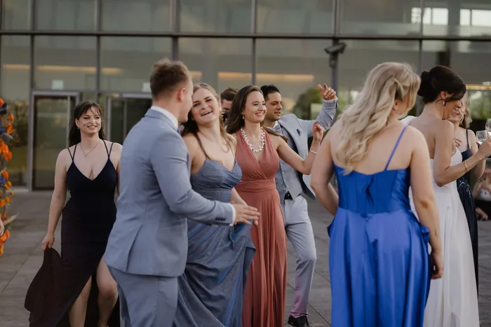 Junge Frauen und Männer in Abendkleidern und hellen Anzügen tanzen lachend vor einer Glasfassade, gestellte Szene aus einem Fotoshooting