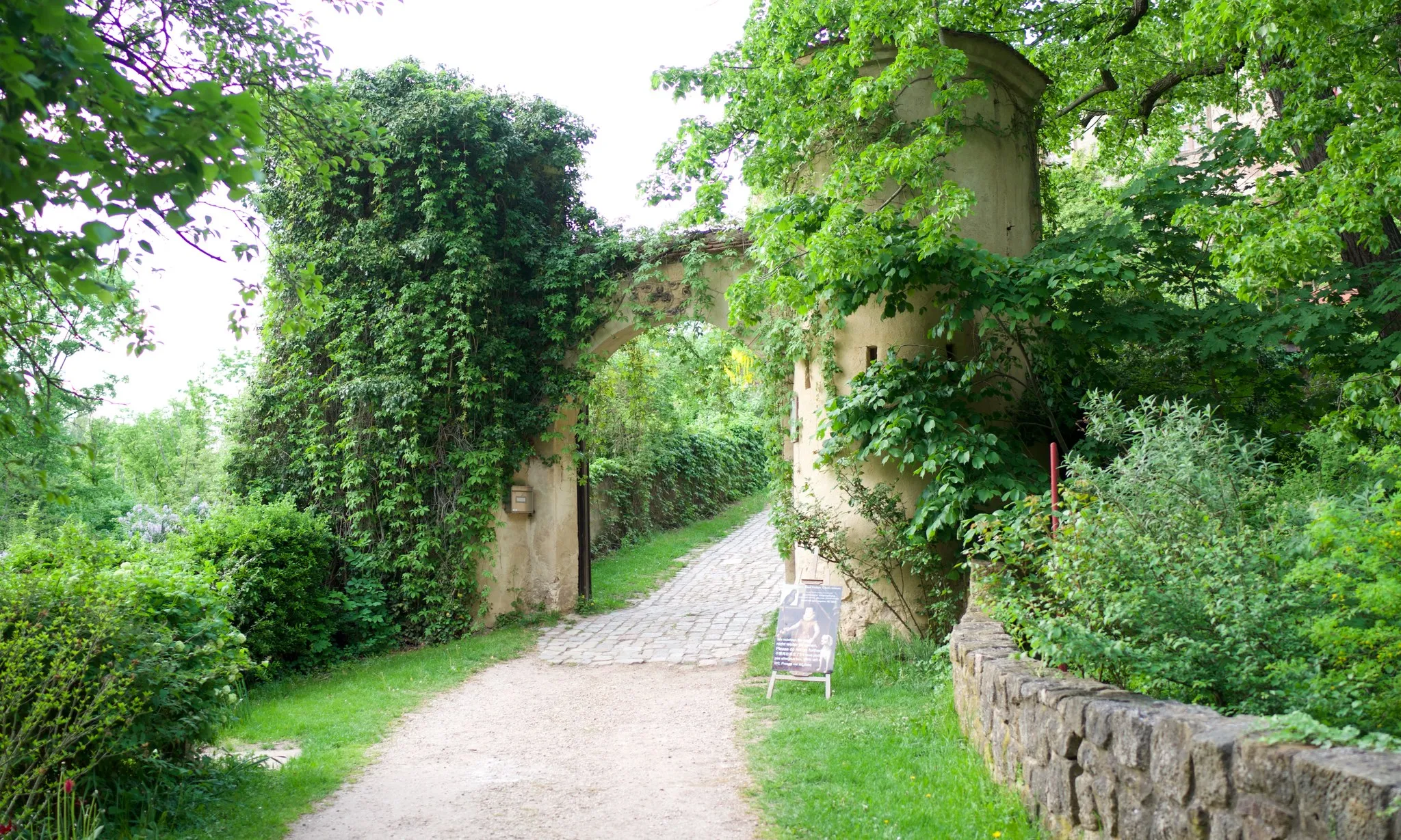 Geschotterter Zufahrtsweg zum Schloss, der durch einen efeuüberwachsenen Torbogen mit Sandsteinrelief führt, rechts daneben ein Rundturm und eine niedrige Bruchsteinmauer